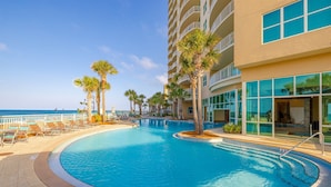 Zona de piscina con sillones reclinables y palmeras, rodeada por un edificio con grandes ventanales y balcones.