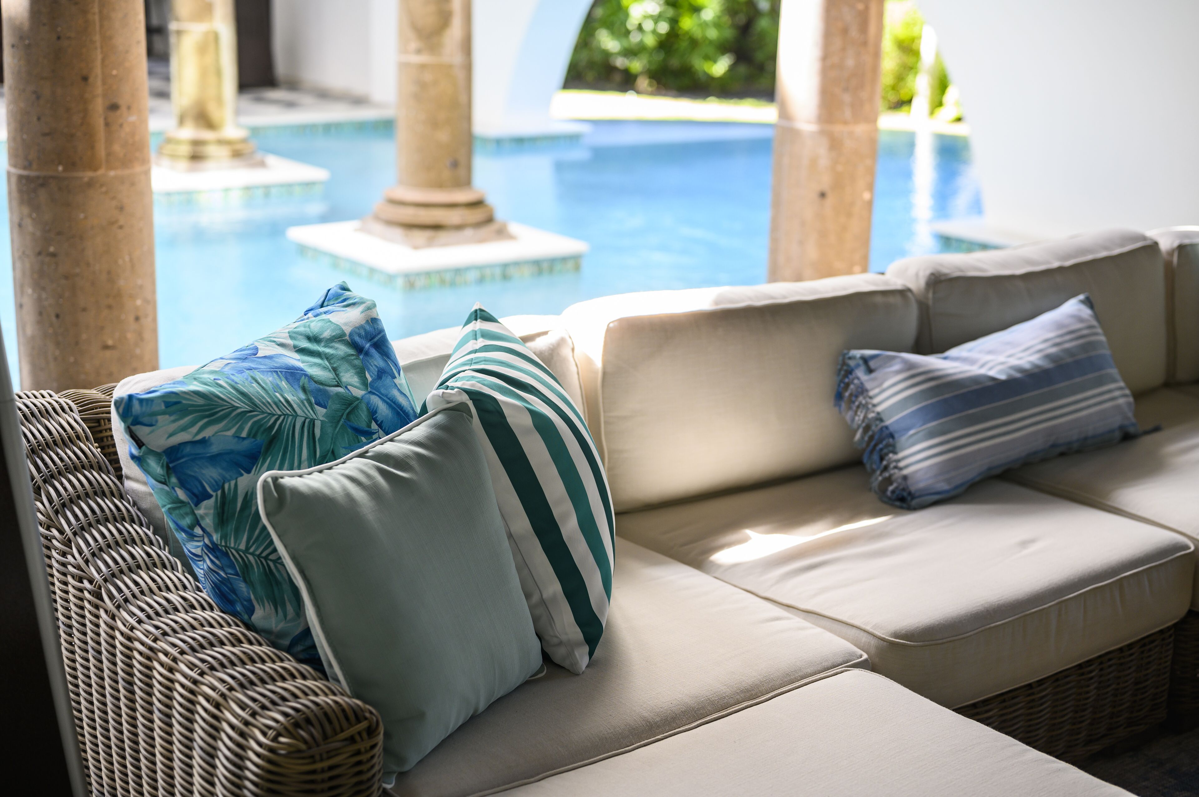A poolside seating area with a wicker sofa and cushions, overlooking a pool with columns and a clear view of the surrounding greenery.
