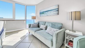 A modern living room with a light blue sofa, a painting of the ocean, and a view of the sea through large windows. - 🌴Gulf Views- Beachside Pool- Studio Top of the Gulf 813🌴 (Panama City Beach)