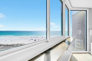 Une vue sur la plage depuis une chambre dotée de grandes fenêtres, d’un banc en bois et d’un mur à la texture particulière.