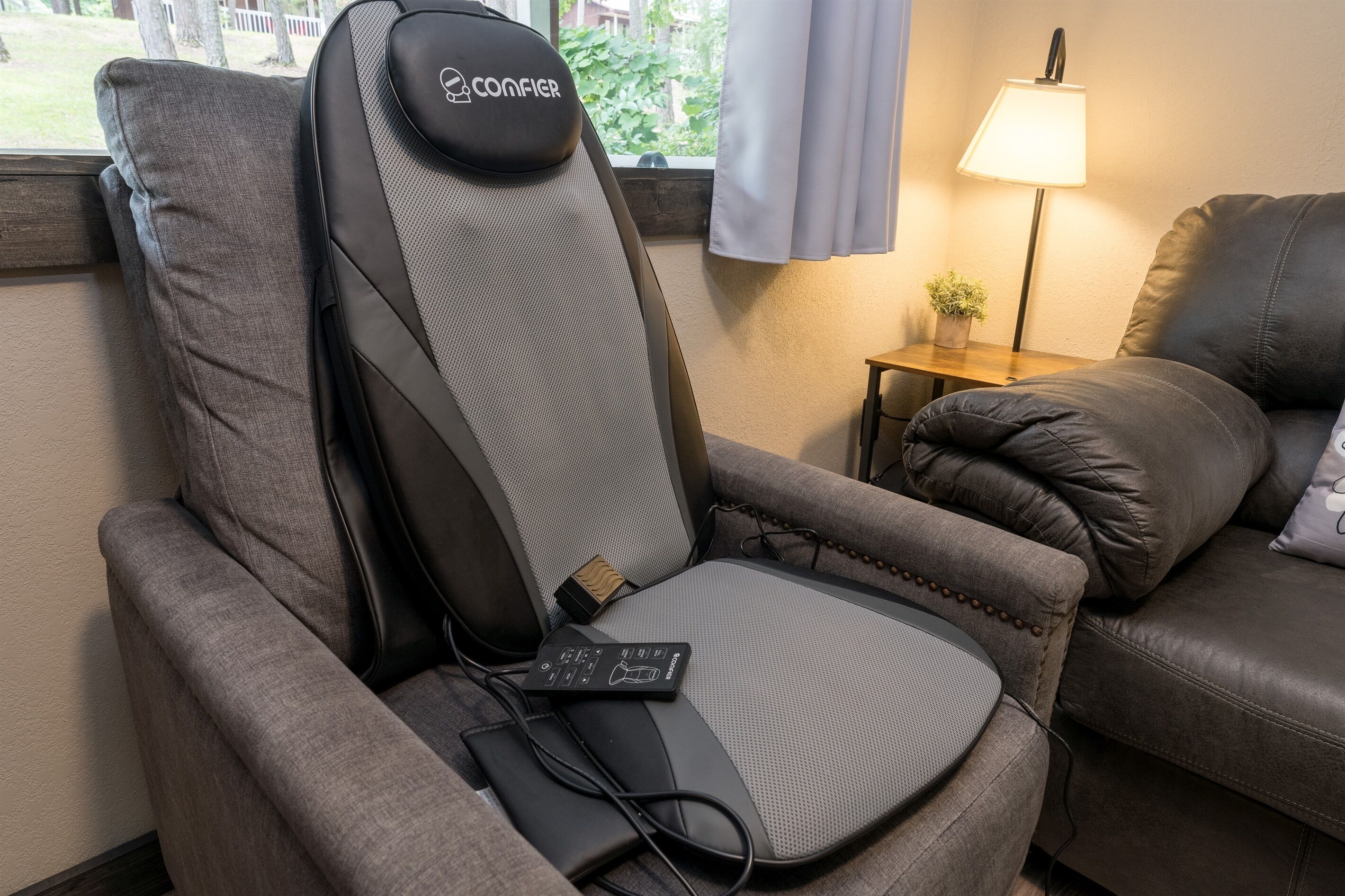 Een grijze massagestoel met bedieningspaneel en oplaadkabel, geplaatst naast een leren bank en een klein tafeltje met een lamp en een plant.