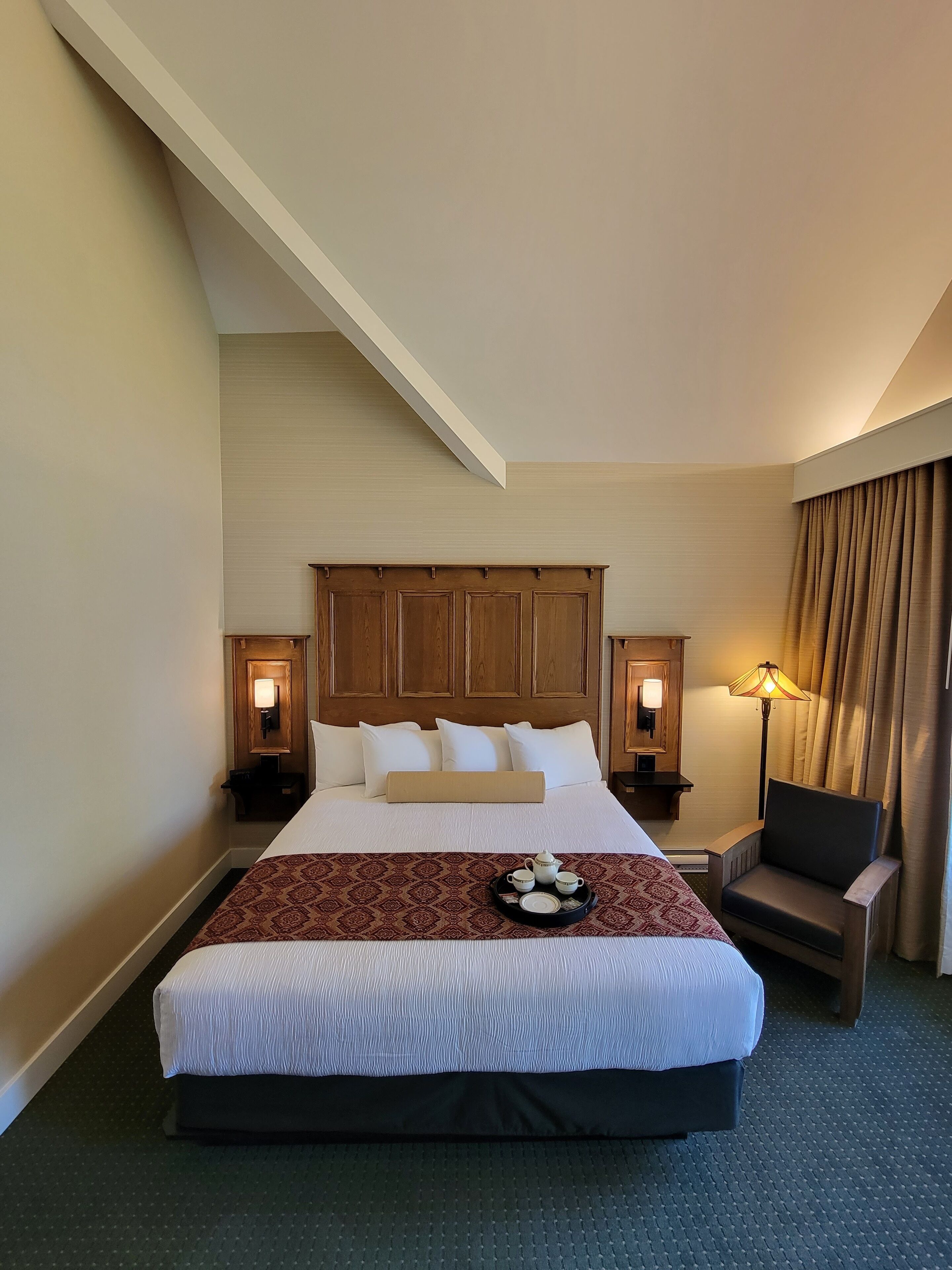 A hotel room with a large bed, two bedside tables with lamps, a chair, and a tray with teapots and cups.