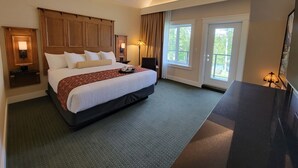 A hotel room with a large bed, a desk, two bedside lamps, a window with curtains, and a door. - Kilmorey Lodge (Waterton Park)