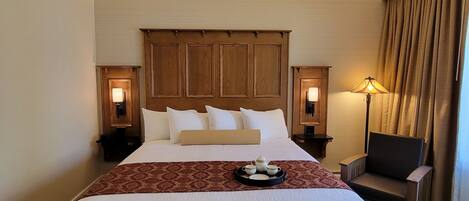 A hotel room with a large bed, a wooden headboard, two bedside lamps, a chair, and a tray with teapots and cups.
