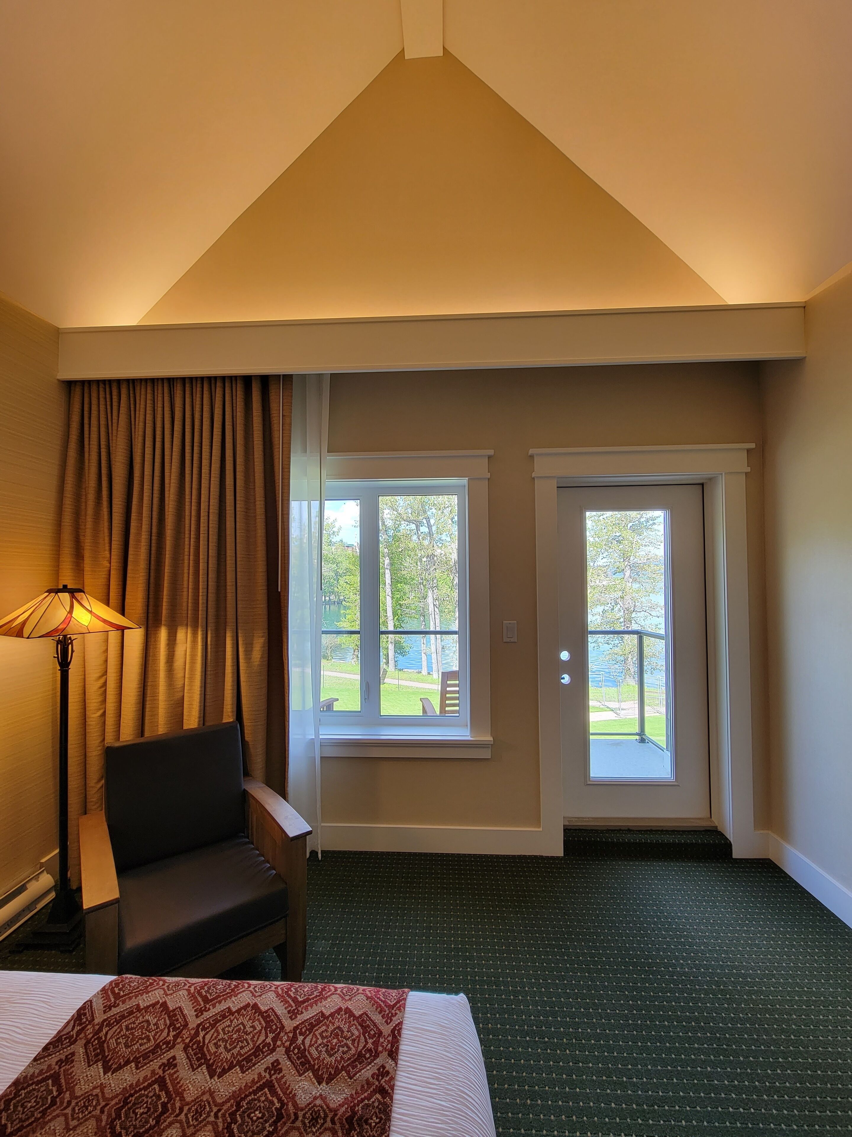 A hotel room with a bed, a chair, a lamp, and two windows with a view of the outdoors.