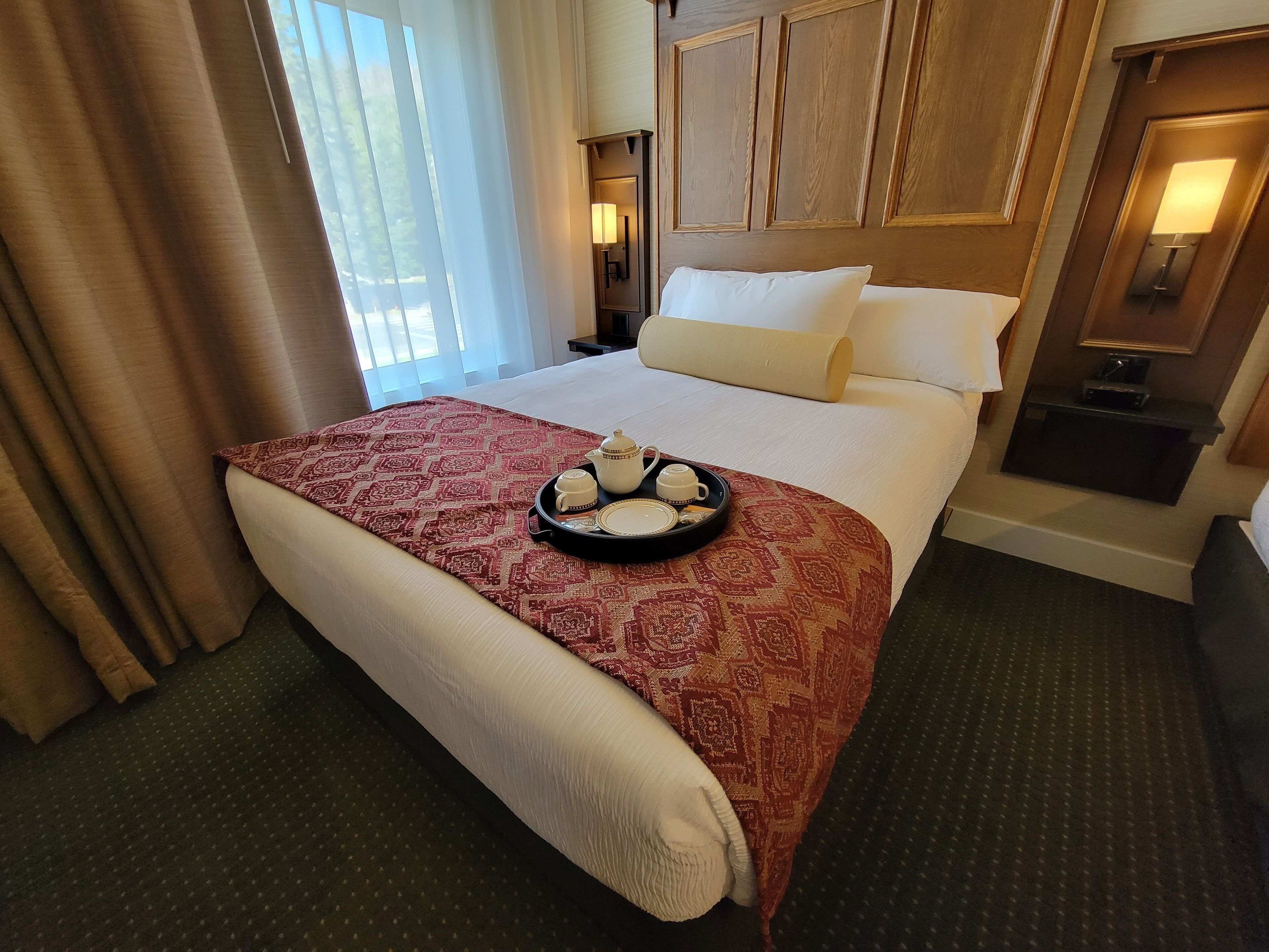 A hotel room with a neatly made bed, a tray with tea and pastries, and a nightstand with a lamp.