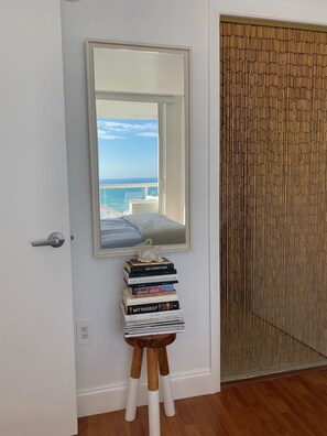 A room with a large mirror reflecting a balcony with a view of the sea, a stack of books on a wooden stool, and a wooden door.