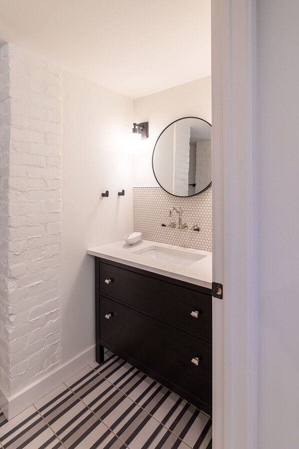 A bathroom with a white brick wall, a white sink with a hexagonal tile backsplash, a round mirror, and a black vanity with drawers.