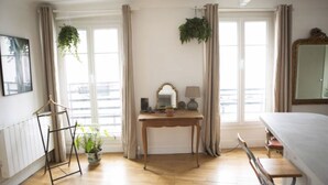 Ein Hotelzimmer mit einem Holztisch, einem Bett, einer hängenden Pflanze und einem großen Fenster mit Vorhängen.