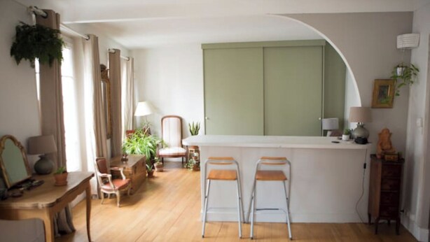 Ein gemütliches Wohnzimmer mit Holzboden, eine Essecke mit Tisch und Stühlen, eine Küche mit Arbeitsfläche und Barhocker und ein großes Fenster mit Vorhängen.