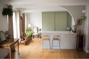 A cozy living room with a wooden floor, a dining area with a table and chairs, a kitchen with a counter and stools, and a large window with curtains.