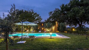 A swimming pool with lounge chairs and an umbrella, surrounded by a well-maintained lawn and trees, with a stone building in the background.