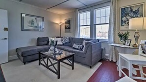 Ein Wohnzimmer mit einem grauen Sofa, einem Couchtisch und einem weißen Tisch. Zwei Fenster mit Vorhängen, eine Lampe und gerahmte Bilder schmücken die Wände.