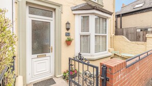 A residential building with a white door, a bay window with shutters, a small outdoor seating area, and a brick wall. - Postman's Cottage (Bridlington)