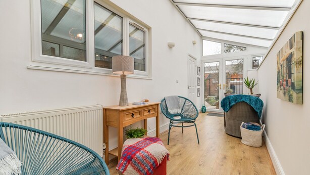 A narrow hallway with a wooden floor, a red ottoman, a blue chair, a wooden table with a lamp, and a window with a view of the outside. - Postman's Cottage (Bridlington)