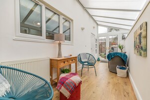 Un pasillo estrecho con suelo de madera, un reposapiés rojo, una silla azul, una mesa de madera con una lámpara y una ventana con vistas al exterior.