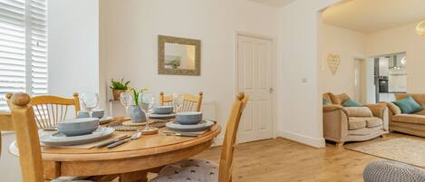 A dining area with a wooden table set for four, surrounded by chairs with patterned cushions. A white door and a heart-shaped decoration are visible on the wall.