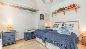 A bedroom with two beds, a bedside table with a lamp, a dresser, and a shelf with decorative items. - Postman's Cottage (Bridlington)