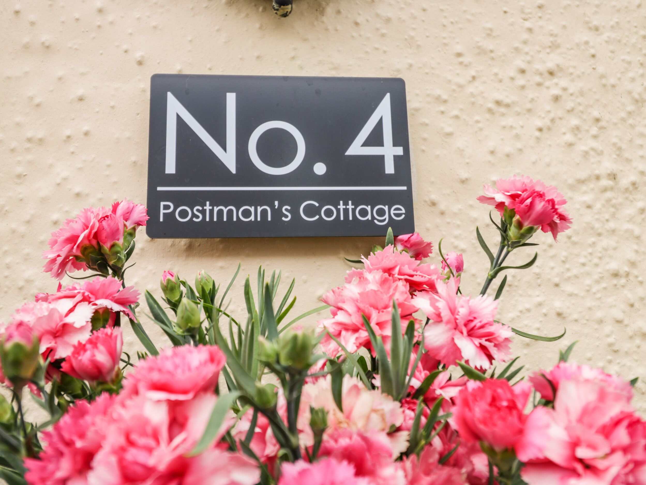 Placa com a inscrição 'No. 4 Postman's Cottage' e flores cor-de-rosa em frente.