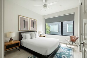 A modern bedroom with a large bed, a ceiling fan, two framed abstract paintings, a nightstand with a lamp, a window with blinds, and a patterned rug. - East Austin Oasis | Pool | Grill | Modern 4BR Home (Austin)