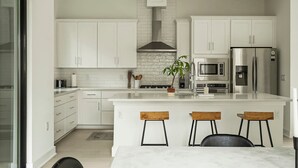 Een moderne keuken met witte kasten, een roestvrijstalen koelkast, een magnetron en een gasfornuis. Er staan drie houten krukken bij het eiland.