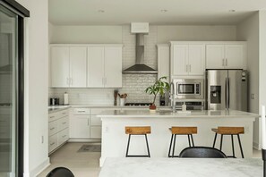 A modern kitchen with white cabinets, a stainless steel refrigerator, a microwave, and a gas stove. There are three wooden stools at the island. - East Austin Oasis / Pool / Grill / Modern 4BR Home (Austin)