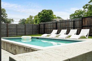 A rectangular swimming pool with a tiled edge, surrounded by a concrete deck. There are four white lounge chairs aligned along the poolside. - East Austin Oasis | Pool | Grill | Modern 4BR Home (Austin)