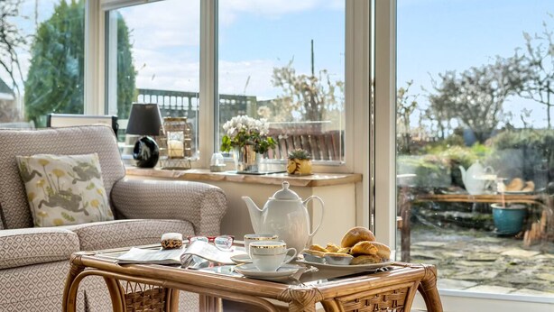 Un salon chaleureux avec une table en osier mise pour le thé, un canapé agrémenté de coussins à motifs et une vue sur un jardin, visible à travers de grandes fenêtres.