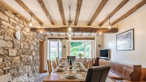 Un espace repas avec une longue table en bois dressée pour le repas, des murs en pierre et une vue sur la verdure à l’extérieur.