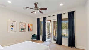 Un dormitorio con cama, ventilador de techo, una silla y puerta corrediza de vidrio que da a un espacio exterior.