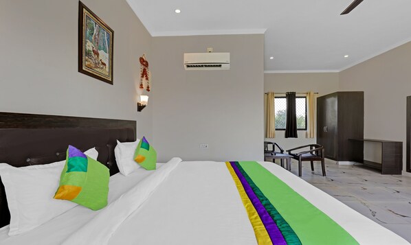 A hotel room with a large bed, a headboard, a wall-mounted air conditioner, a painting, a wall-mounted light fixture, and a window with curtains.