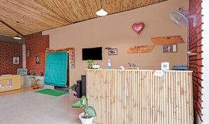 A reception area with a bamboo reception desk, a green mat, potted plants, a heart-shaped balloon, and a sign indicating check-in/out times.