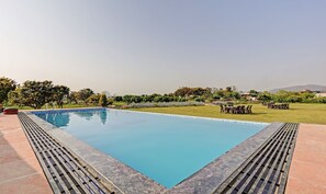 Una piscina rectangular con borde de baldosas, rodeada de césped, árboles y una valla.