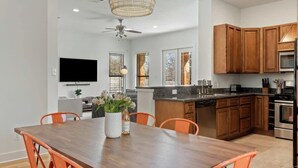 A modern kitchen with wooden cabinets, a dining area with a wooden table and orange chairs, a ceiling fan, and a television mounted on the wall. - Bright 3BR Home / East ATX / Cowboy Pool / Near UT (Austin)