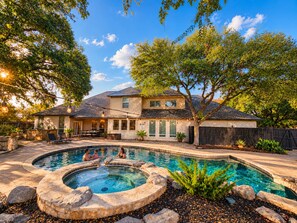 Une maison à deux étages avec une piscine dans le jardin, entourée d’arbres et dotée d’une terrasse en pierre.