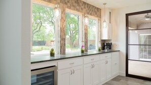 Una cocina con armarios blancos, una encimera y luces colgantes. Hay ventanas con vistas a árboles y un patio.