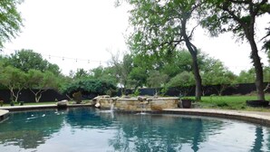 Piscina con cascada de piedra, rodeada de un jardín ajardinado con árboles y un muro de piedra.