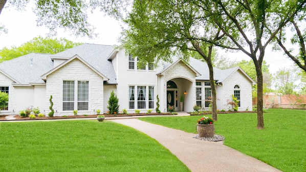 A white two-story house with a stone facade, a front entrance with an arch, and a well-maintained lawn with a pathway leading to the door.