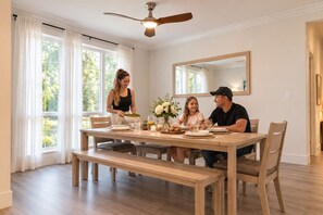 Une salle à manger avec une table en bois, des chaises, un ventilateur de plafond et une grande fenêtre avec des rideaux.