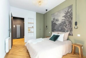 Une chambre à coucher comprenant un lit, une table de chevet, une lampe fixée au mur, un radiateur et une porte donnant sur une autre pièce.