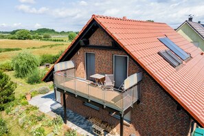 A two-story house with a red-tiled roof, a balcony with a glass railing, and a small table and chair.