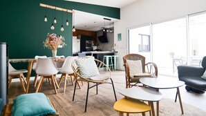 A modern living room with a dining area, featuring wicker furniture, a large window, and a kitchen in the background. - Beautiful holiday home 7 minutes from the beach in Brem sur Mer (Brem-sur-Mer)