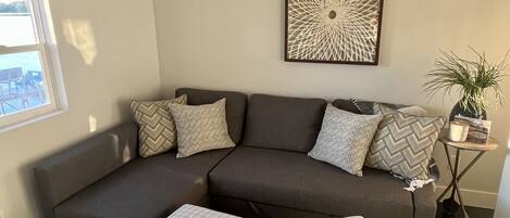 Ein modernes Wohnzimmer mit einem Ecksofa, zwei Hocker, einem gemusterten Teppich, einer Zimmerpflanze und einem Wandbild.