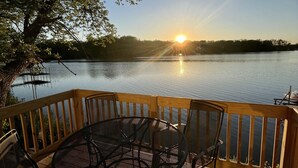 A wooden deck with a round table and chairs overlooking a calm lake at sunset.