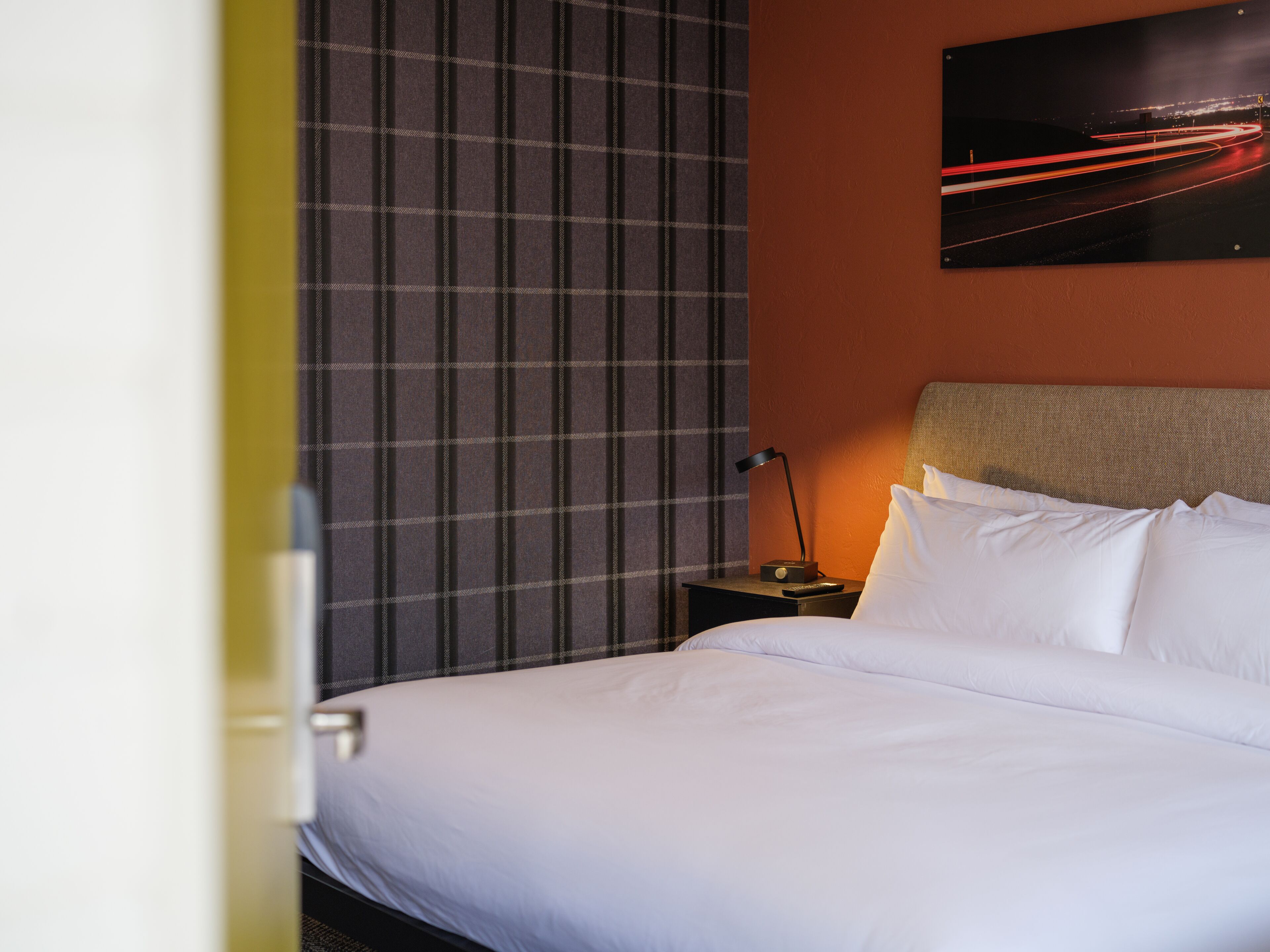A hotel room with a bed, a nightstand, a lamp, and a painting of a cityscape.