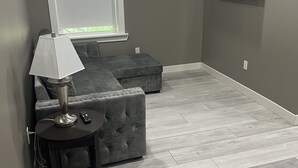 A room with grey walls, a grey sectional sofa, a small round table with a lamp, and a TV mounted on the wall.