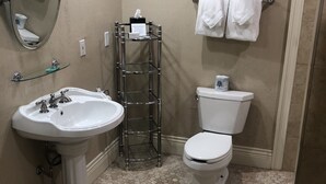 A bathroom with a pedestal sink, toilet, and a mirror. There is a shelf with a tissue dispenser and a trash can. - Niagara Crossing Hotel and Spa King Room (Lewiston)