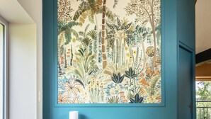 Una habitación con un amplio mural de temática tropical en la pared, un sillón gris y una mesa de madera con una lámpara.