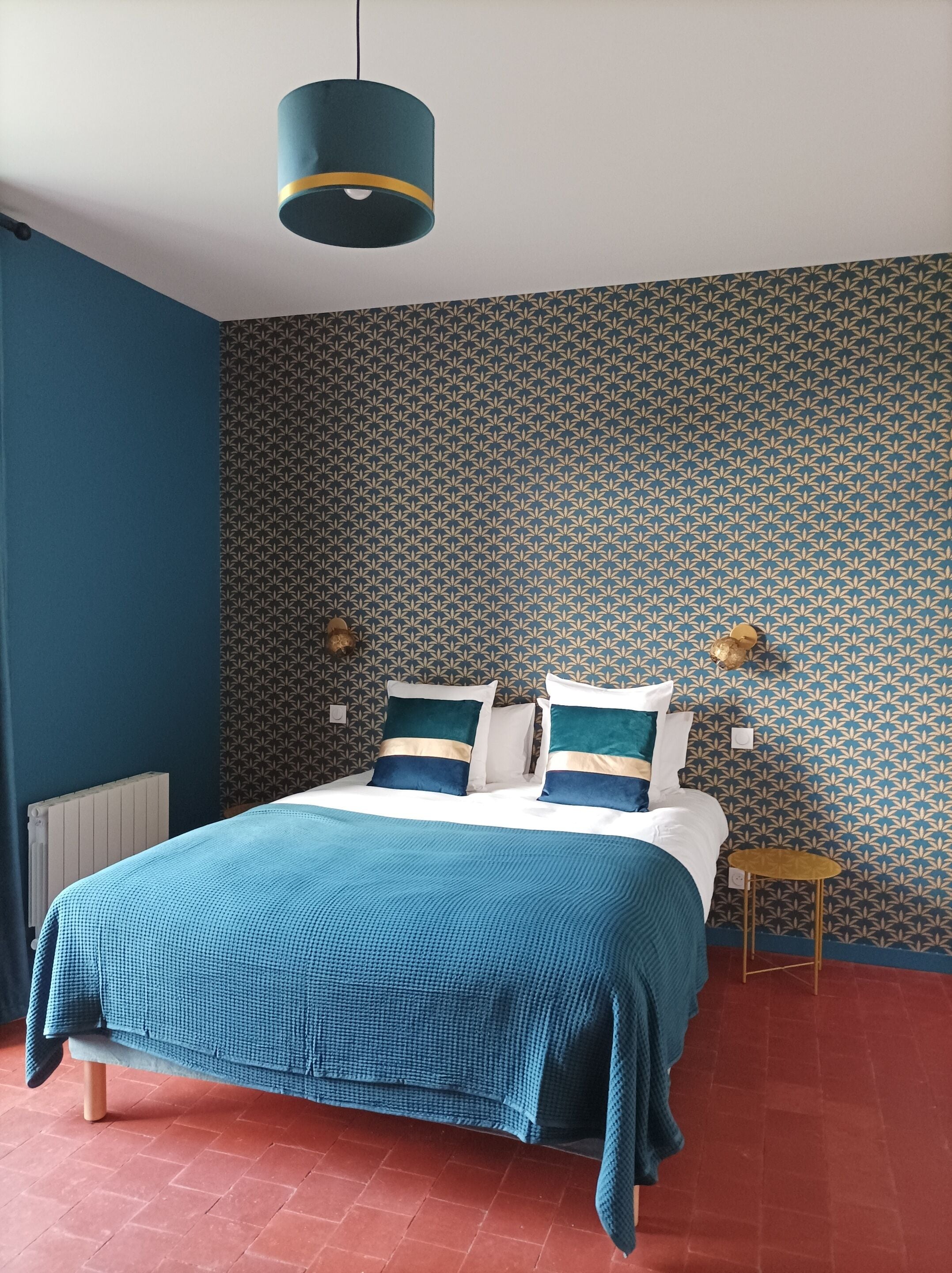 Une chambre à coucher avec un lit, des murs bleus, un papier peint à motifs, une petite table de chevet et un luminaire suspendu.