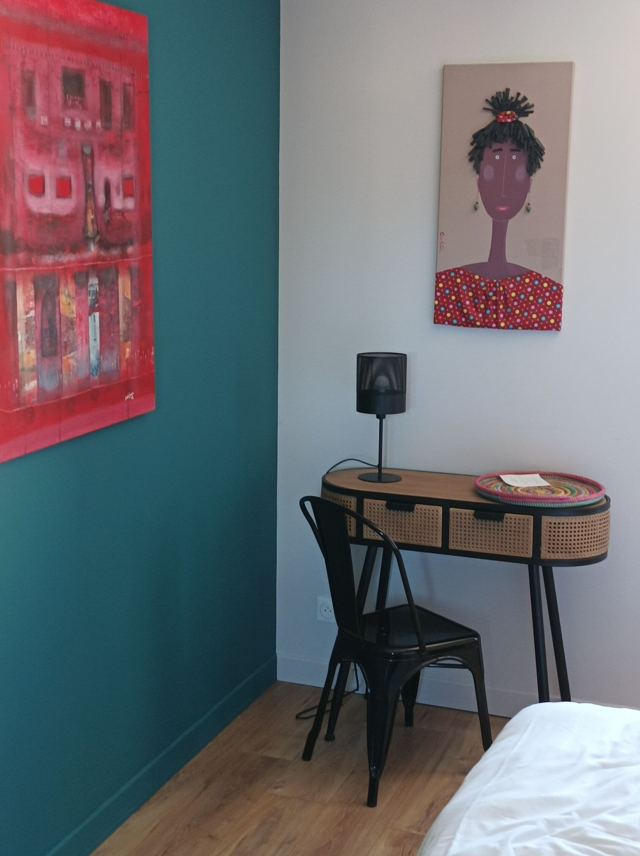 A room with teal walls, a wooden desk with a chair, a bed with white bedding, a red abstract painting, and a portrait on the wall.
